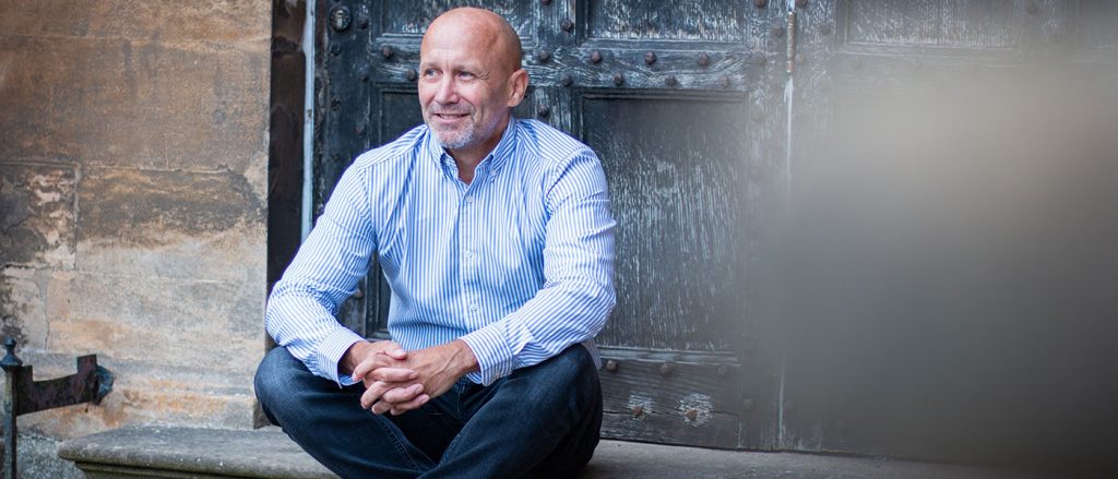 Bald male sat in a doorway, wearing a stripey shirt, smiling and looking into the distance