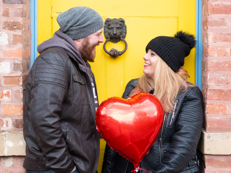 A couple stood in front of a yellow door holding a red love heart balloon