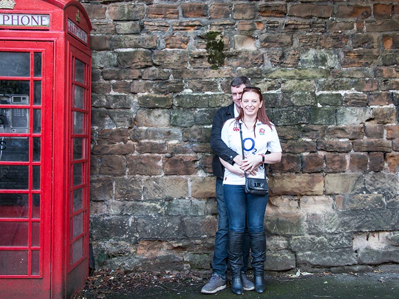 Couple cuddling against a brick wall next to a red telephone box