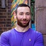 Dating profile photo of a man wearing a bright purple t-shirt, smiling and leaning into a wall in Nottingham