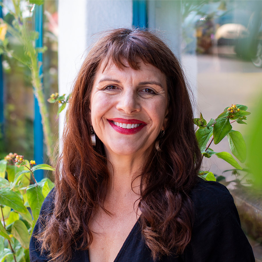 Dating Photo of a female with long brunette hair, wearing bright red lipstick. Sat on a bench outside a shop surrounded by plants.