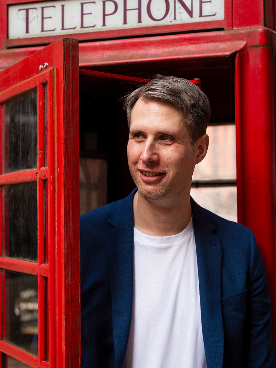 Male stood in a telephone box wearing a white t-shirt and navy blazer