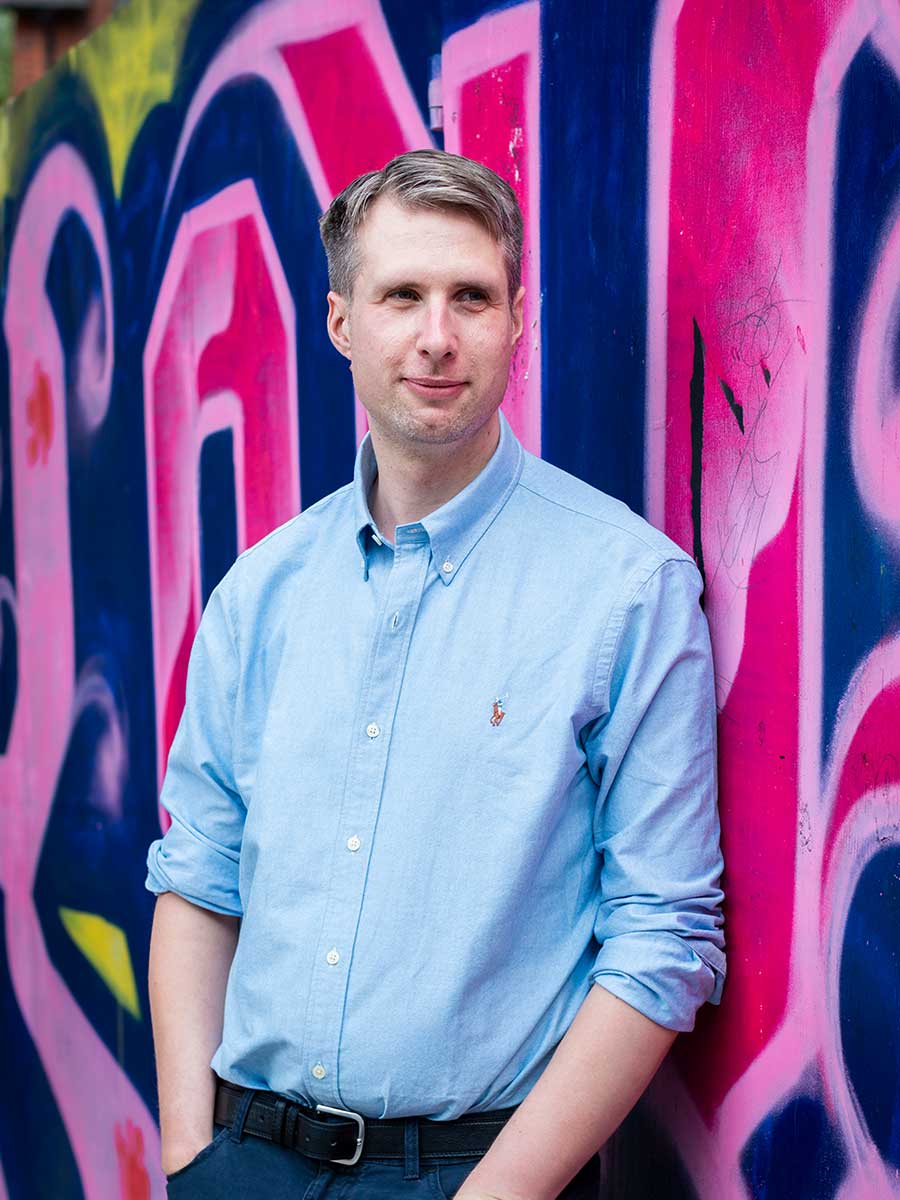 Single man stood against a brightly coloured graffiti wall. He wears a pale blue denim-style shirt and dark jeans.