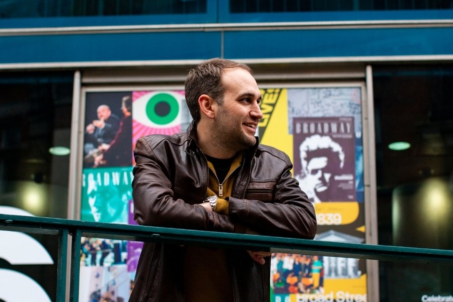 A guy leaning on a railing outside a cinema, he wears a brown leather jacket and looks to his left, smiling.