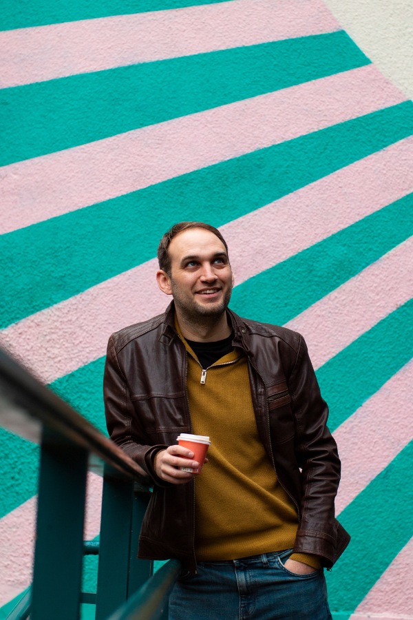 A guy leaning on a railing with a colourful mural in the background. He wears a mustard jumper and brown leather jacket.