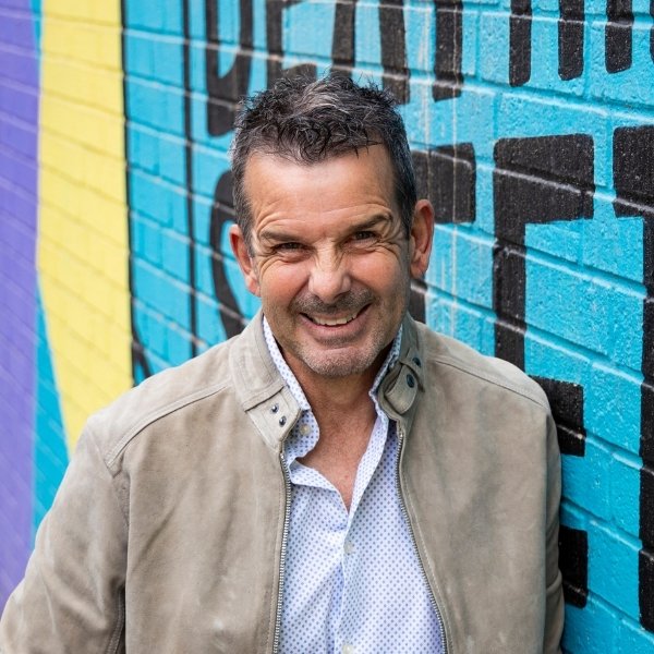 A male client leaning against a wall with a bright mural painted on. He wears a white patterned shirt with a beige open jacket, and has a lovely big smile towards the camera. This dating photo was captured in Nottingham City Centre.